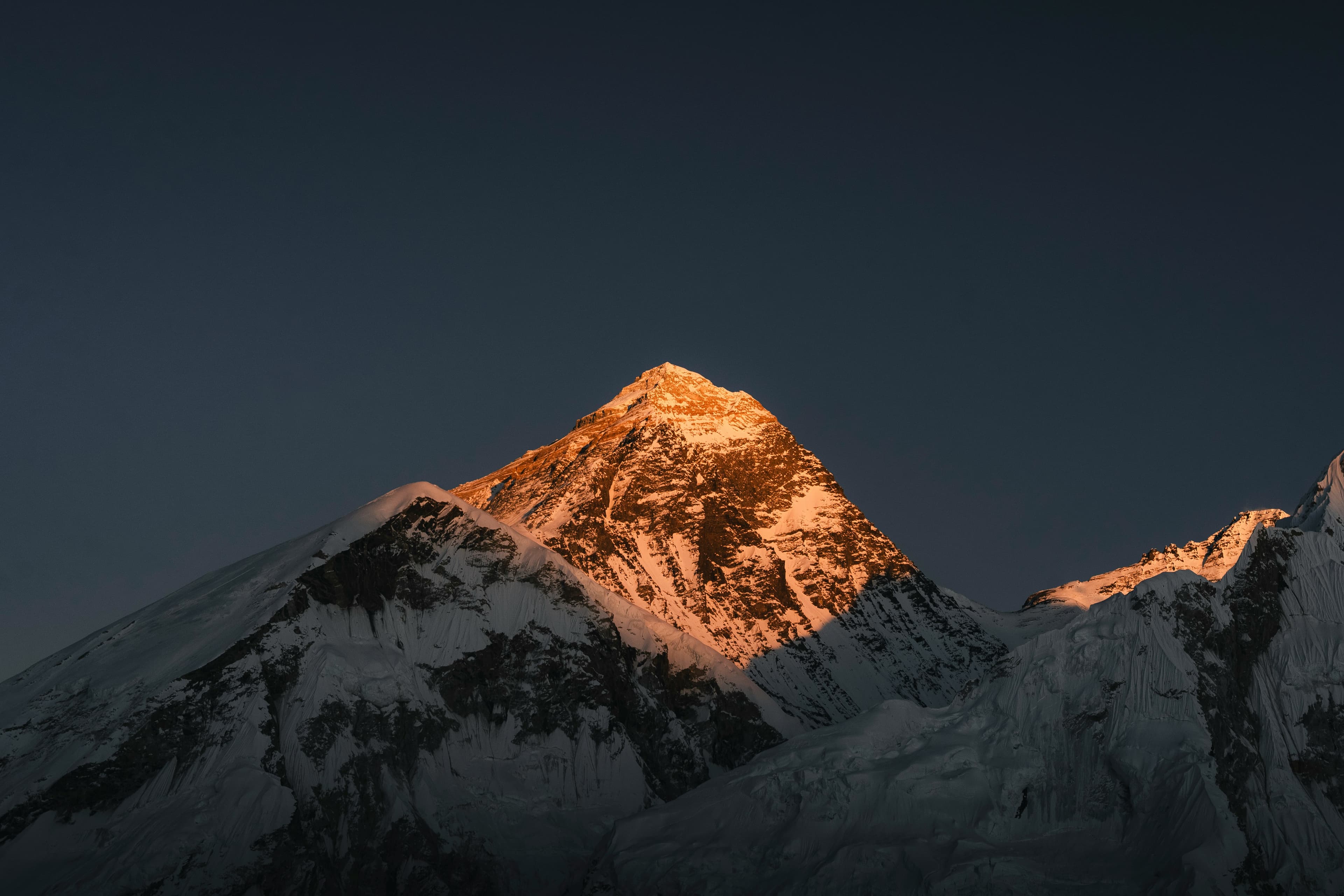 Two trekkers with backpacks walking a gravel trail past tall prayer flags and dry stone walls stacked with firewood, beneath a sunlit cliff in a high Himalayan village