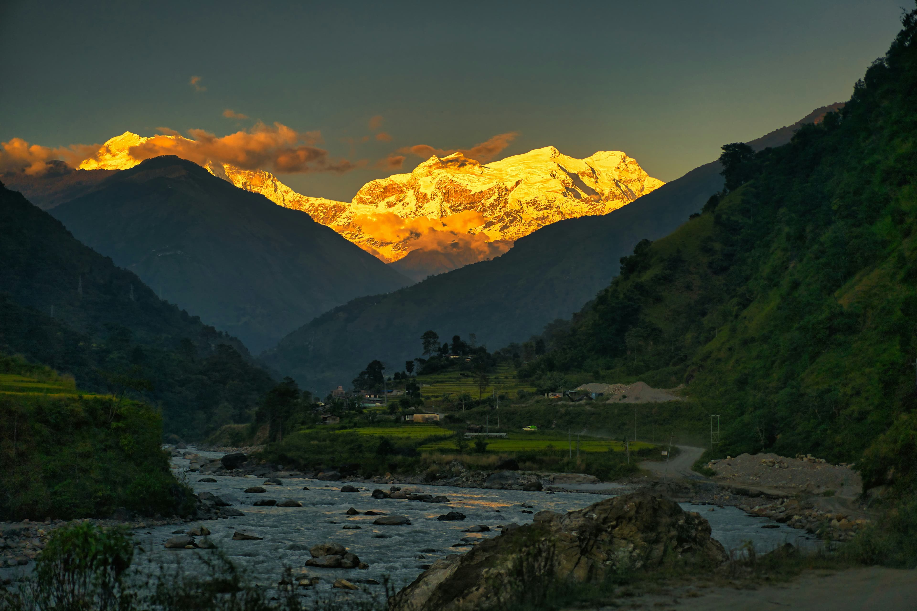 Golden dusk light on a snow-capped Himalayan peak above a rocky river valley, with terraced fields and a small hillside village